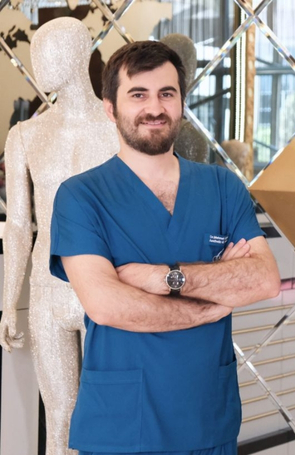 A person with a beard stands confidently with arms crossed, wearing blue scrubs and a wristwatch. They are a skilled surgeon specializing in necklifts, positioned in front of a sparkling mannequin, with a decoratively mirrored wall behind them.